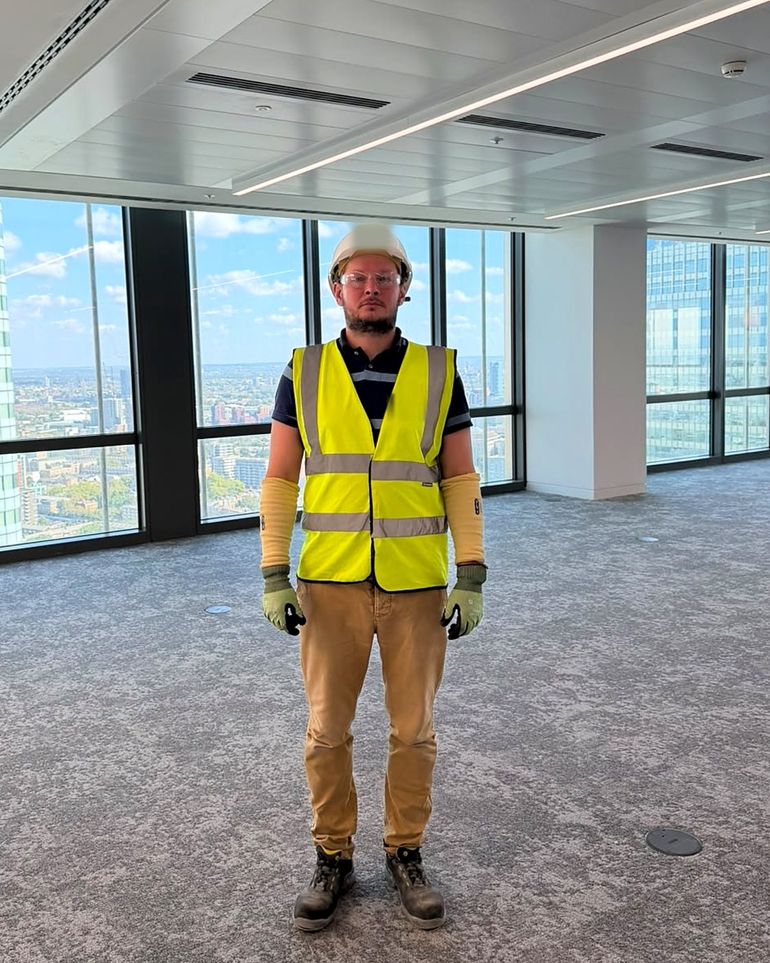 Vitalii Kuziutyn in full site PPE - hard hat, safety glasses, hi-vis vest, cut-resistant sleeves and gloves - standing on a finished office floor beneath a metal plank ceiling.